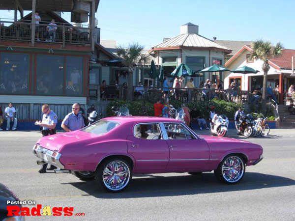 A crowd watches as a pink car cruises down the street.