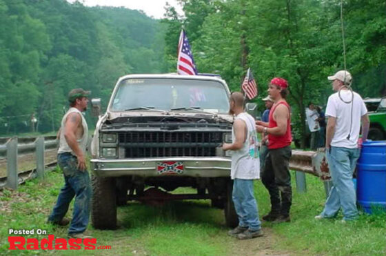 A group of rednecks standing around a truck on a dirt road.