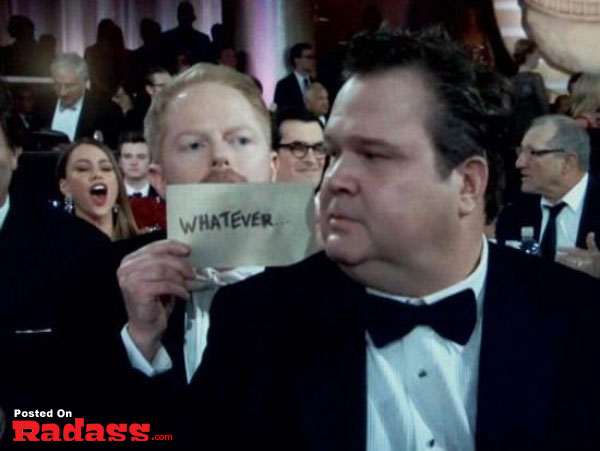 A man in a tuxedo photobombs a celebrity-style event while holding up a sign.