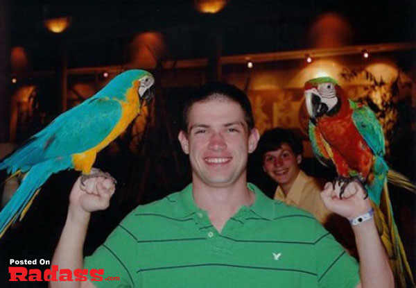 A man photobombs celebrity-style while holding two parrots in front of him.