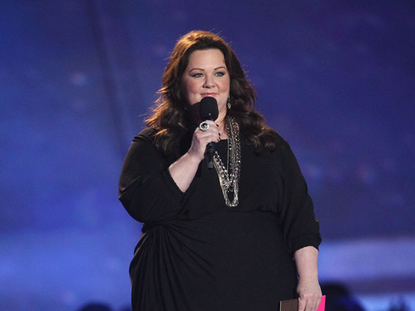 Melissa McCarthy holding a microphone in a black dress.
