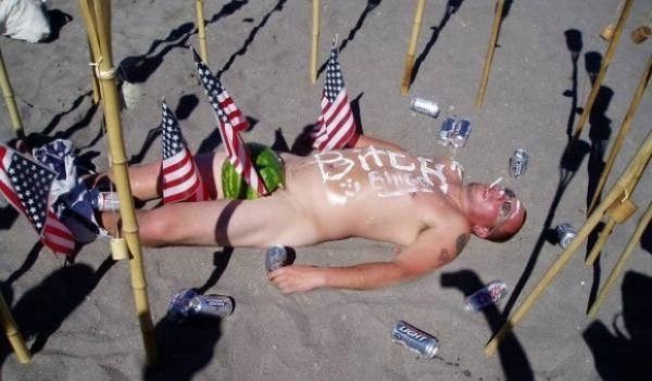 A man, intoxicated and surrounded by friends, rests on the sand with an American flag painted on his face.
