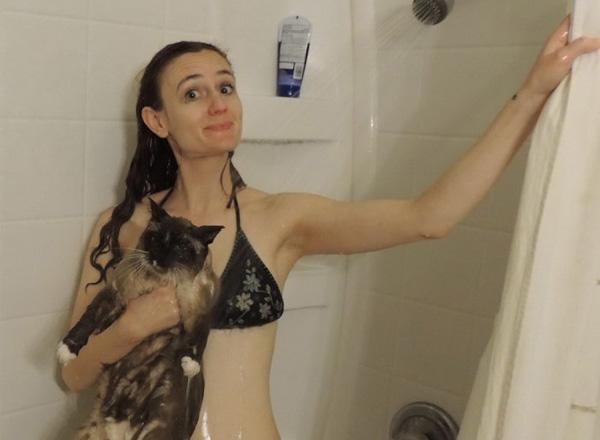 A woman in a bikini playfully holding a cat in the shower, expressing 