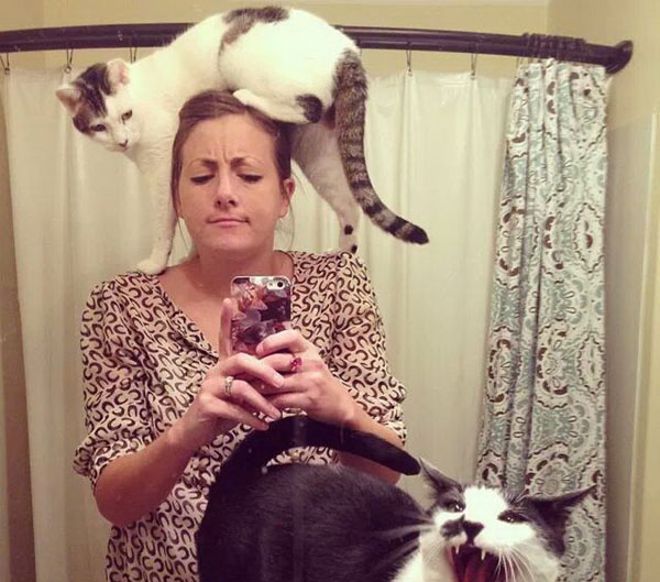 A woman taking a selfie with two cats on her head.