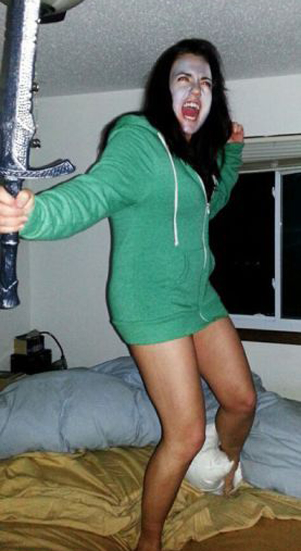 A woman in a green hoodie wielding a sword on a bed, showcasing her fierce and enigmatic persona.