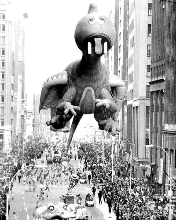 A large dinosaur balloon flying over a city street during Macy's Day Parade.