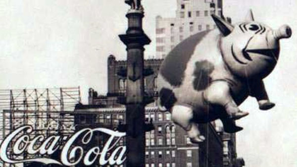 An old photo of a coca cola balloon flying over a city during Macy's Day Parade.