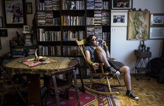 A man sitting in a rocking chair in a room with bookshelves and a table, offering Another View On Iran.
