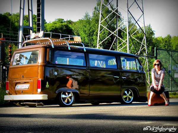 A woman is posing with a vintage brown VW bus, showcasing both babes and dubs.
