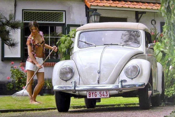 A woman is sweeping a lawn with a VW Beetle, filled with babes.