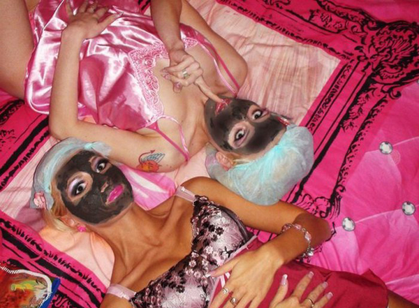 Two women laying on a pink bed with black masks on their faces, continuing the 