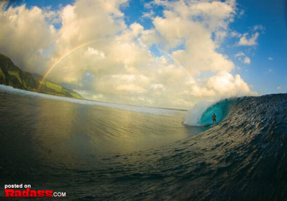 A surfer is riding a wave with a rainbow in the background. Captured in an inspirational photograph.