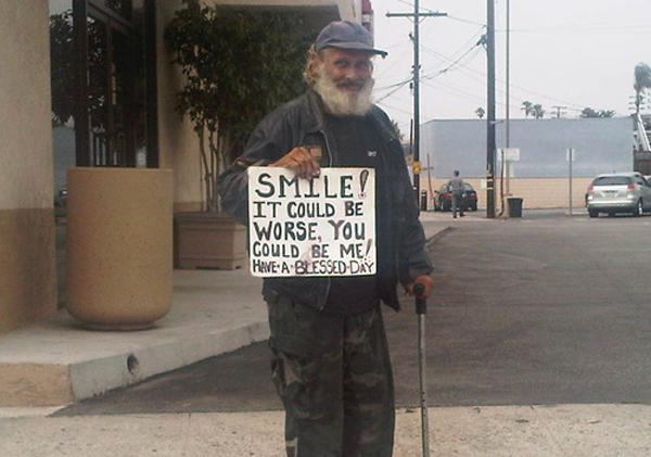 An old man holding a sign that says smile, reminding us that Things Could Be Worse.
