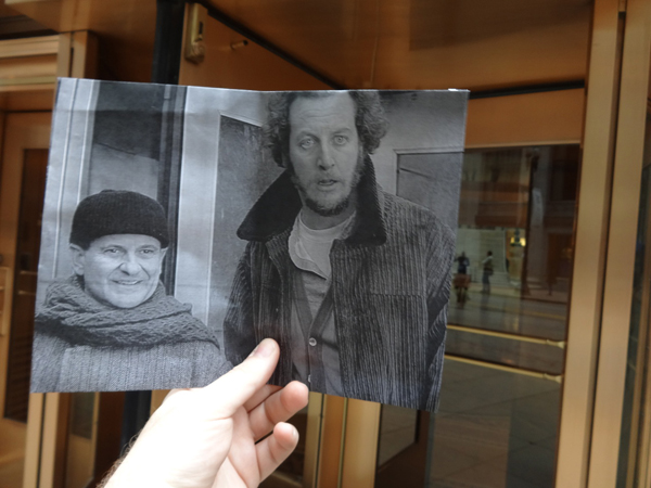 A photographer matching movie scenes with present-day locations, captures and holds a photo of two men in front of a building.