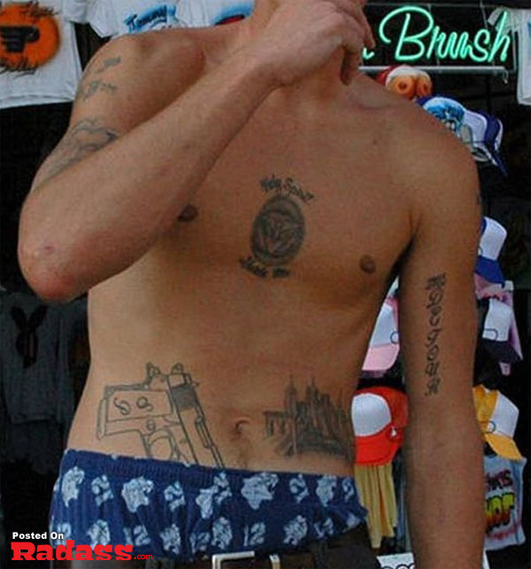 A tattooed man poses shirtless in front of a store.
