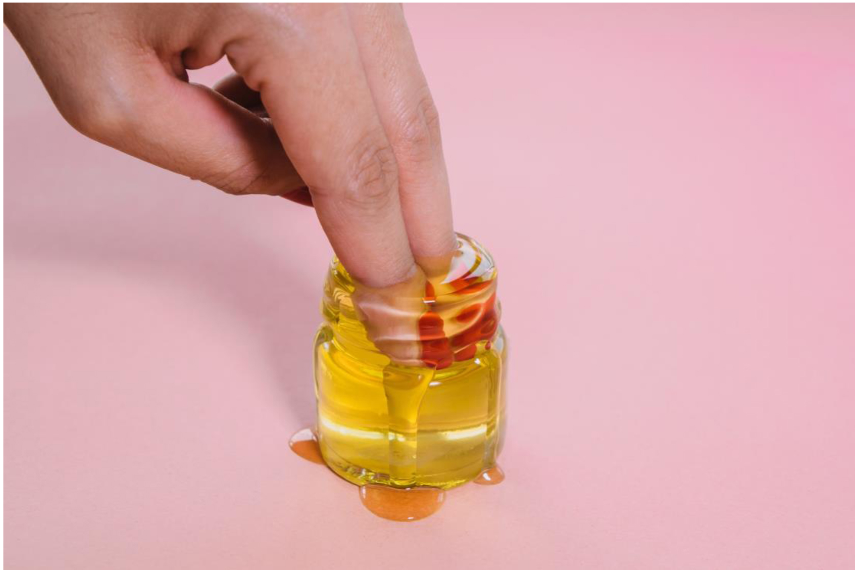 A hand holding a small bottle of honey on a pink background featuring genital tunes.