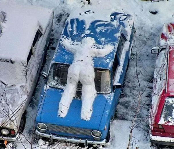 A blue car covered in snow with a snowman on top, showcasing the snowy winter scenery.