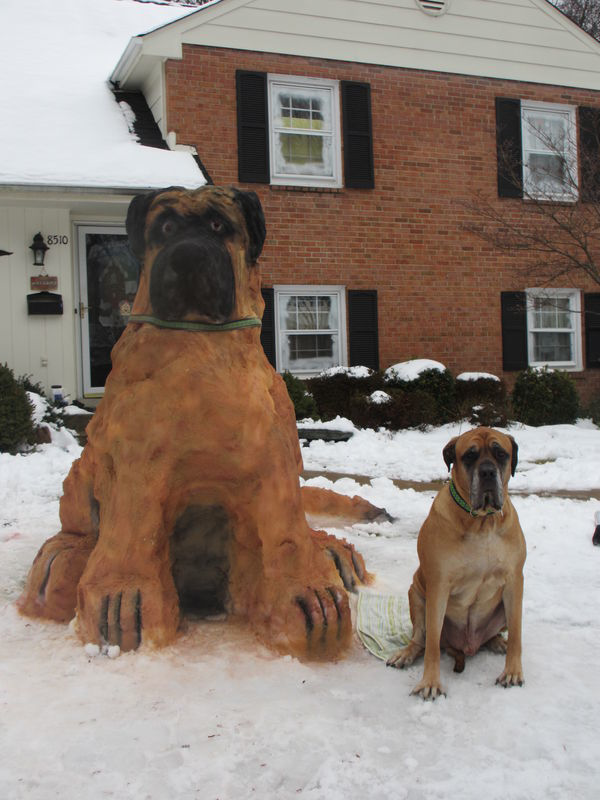 A dog statue sitting in front of a snowy house.